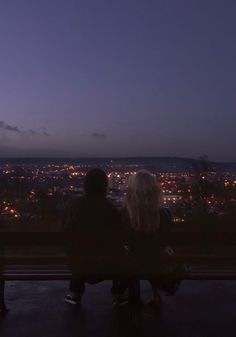 two people are sitting on a bench looking at the city lights in the night sky