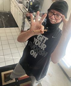 a man in a kitchen making the peace sign with his hand and wearing a black t - shirt that says get your nose on