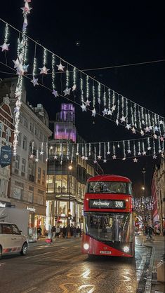 a red double decker bus driving down a street next to tall buildings with christmas lights