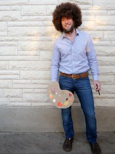 a man with an afro holding a paintbrush and palette in front of a brick wall