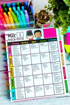 a weekly planner with markers and pens on a white wooden table next to a potted plant