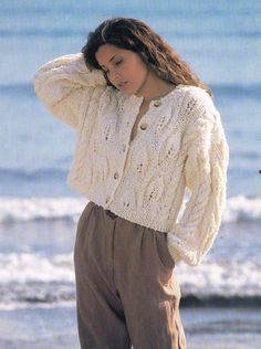 a woman standing on top of a beach next to the ocean wearing a white sweater and brown pants