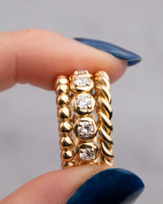 A close-up image of a hand holding a set of gold stacking rings. The rings feature a mix of designs, including polished gold beads, a twisted rope texture, and sparkling diamonds set in rounded bezels. The bold and luxurious textures create a sophisticated and versatile jewelry piece.