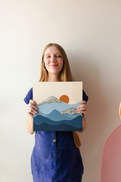 a woman holding up a piece of paper with mountains and waves on it in front of a white wall