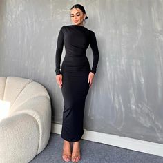 a woman standing in front of a wall wearing a long black dress with an asymmetrical neckline