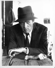 a man in a suit and hat leaning on a table with his hands folded out