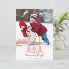 a christmas card with two children on sled in the snow, one is wearing a red jacket