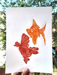 a hand holding up a piece of paper with two goldfish on it and one red fish in the background