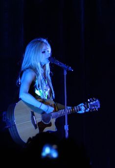 a woman with long blonde hair playing an acoustic guitar in front of a microphone on stage