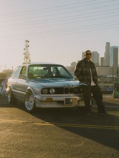 a man standing next to a parked car in front of a tall building with the city skyline behind him