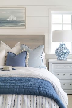 a bed with blue and white pillows on top of it next to a nightstand in front of a window