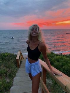 a woman standing on a wooden railing next to the ocean at sunset with her arms behind her back