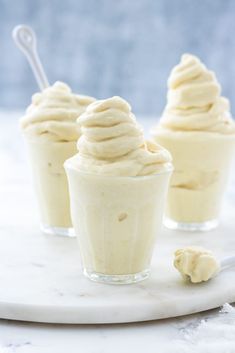 three small cups filled with whipped cream on top of a white marble plate next to two spoons