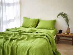 a bed with green sheets and pillows next to a plant on a table in front of a window