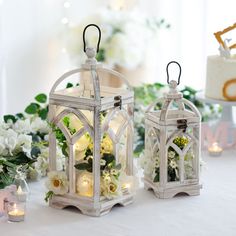 two small white lanterns with flowers and candles on a table next to eachother