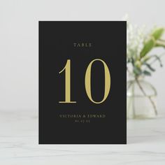 a black and gold table number card with the number ten printed on it, sitting next to a vase filled with flowers