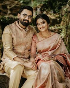 a man and woman sitting next to each other in front of some trees, smiling at the camera