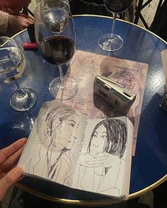 a person is holding up a book with drawings on it next to some wine glasses