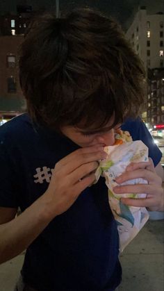 a young man is eating a sandwich on the sidewalk at night in front of some tall buildings