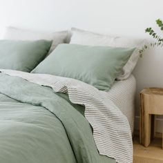 an unmade bed with light green sheets and pillows in a white room next to a small wooden side table