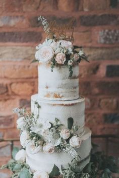 a three tiered cake with flowers and greenery on the top is sitting in front of a brick wall