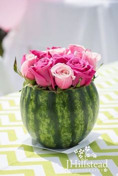 a watermelon vase filled with pink roses on top of a green table cloth