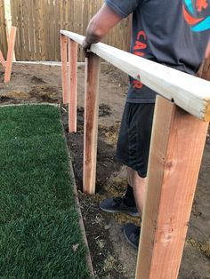 a man standing next to a wooden fence with grass growing on it and another person holding a piece of wood in the background