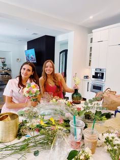 two women sitting at a table with flowers and vases in front of them on the counter