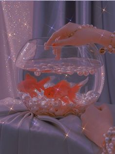 a woman's hand reaching for goldfish in a bowl filled with water and pearls