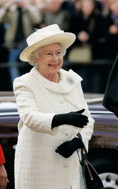 the queen of england is wearing a white coat and black gloves as she stands in front of a car