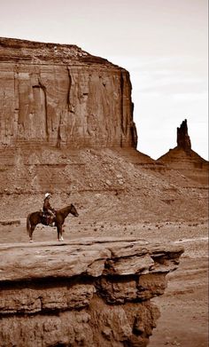 a man riding on the back of a brown horse across a desert field next to a cliff