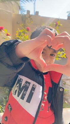 a young boy making a heart with his hands while standing in front of some flowers