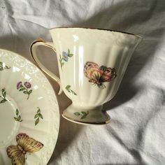 two tea cups and saucers with butterflies on them sitting on a white cloth covered tablecloth