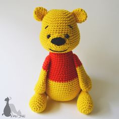 a crocheted yellow teddy bear wearing a red shirt and standing on a white surface