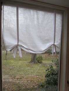 an open window with white curtains on the outside and trees in the yard behind it