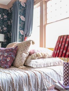 a bed with many pillows on top of it in front of a window next to a lamp