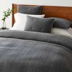 a bed with grey and white comforter next to a vase filled with green plants