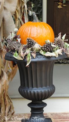 a pumpkin sitting on top of a black urn filled with leaves and pine cones