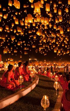 many people are sitting on the ground with lanterns floating in the air