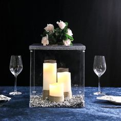 three lit candles in a clear box on a blue table cloth with flowers and wine glasses