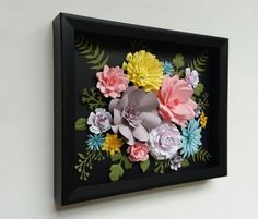 a black frame with paper flowers in it on the wall next to a white wall