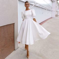 a woman in a white dress standing next to a wall and looking at the camera