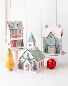 three small christmas houses are sitting on a cake stand and one has a red ornament