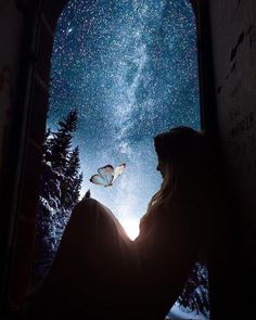 a woman sitting in front of a window looking up at the sky with a butterfly flying over her head