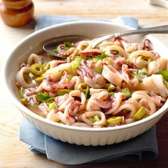 a white bowl filled with pasta and meat on top of a wooden table next to a spoon