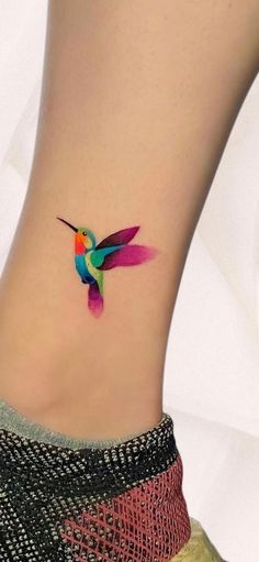 a colorful hummingbird tattoo on the side of a woman's ankle, with pink and green colors