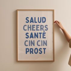 a person holding up a framed poster with the words salud cheers sante cin cin prost