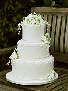 a three tiered white wedding cake with flowers on the top and sides, sitting on a wooden table