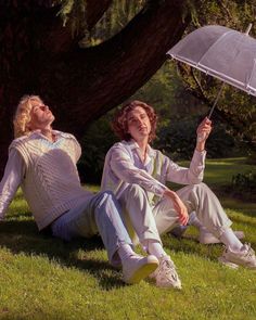 two people sitting on the grass with an umbrella in their hands and one holding another