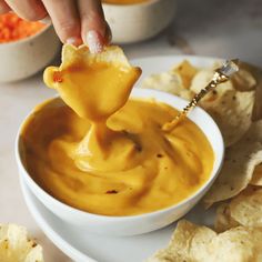 a person dipping tortilla chips into a white bowl filled with cheese dip sauce